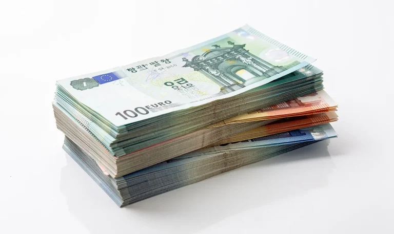 Des liasses de billets sud-coréens et européens sont posées côte à côte sur une table blanche avec une lumière vive et une légère réflexion.