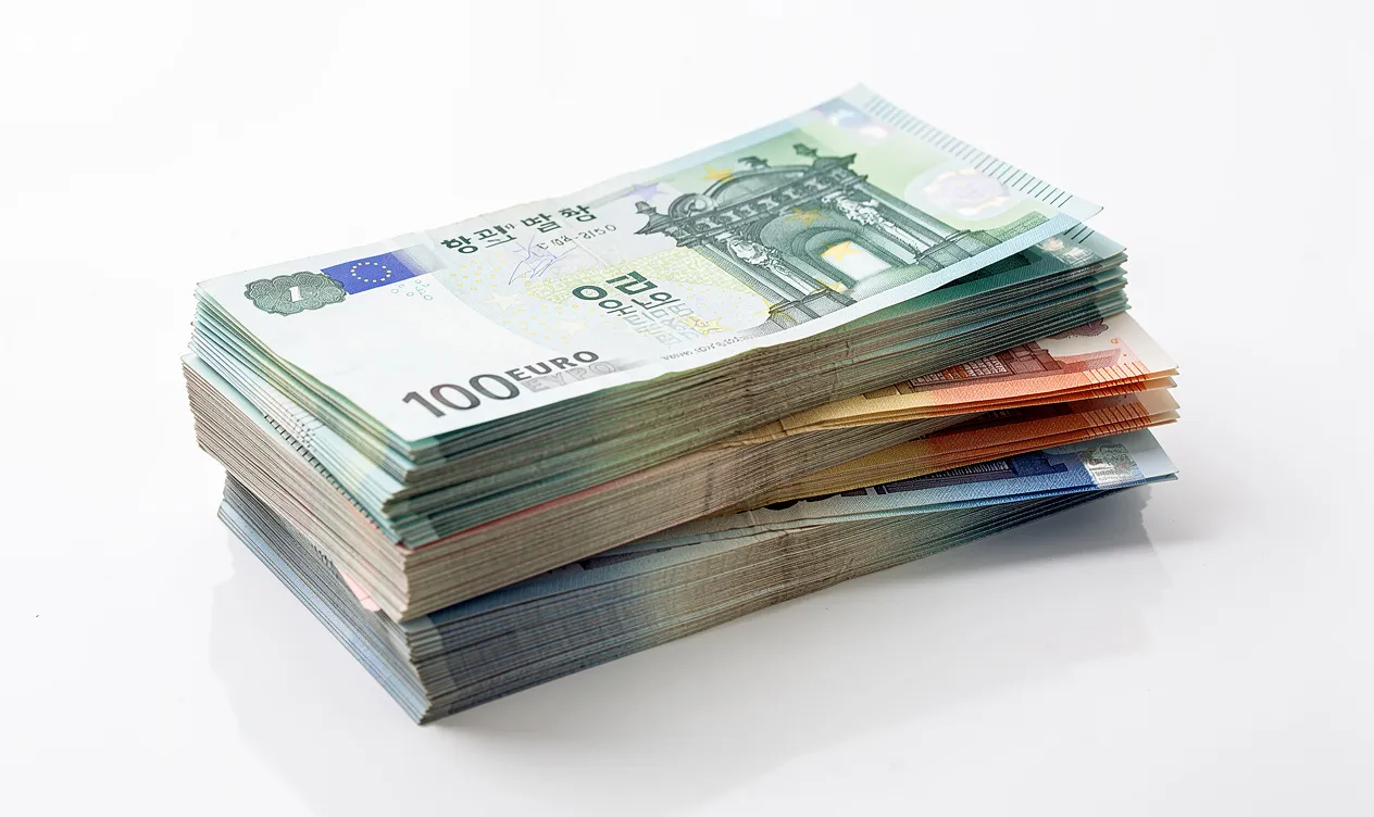 Des liasses de billets sud-coréens et européens sont posées côte à côte sur une table blanche avec une lumière vive et une légère réflexion.