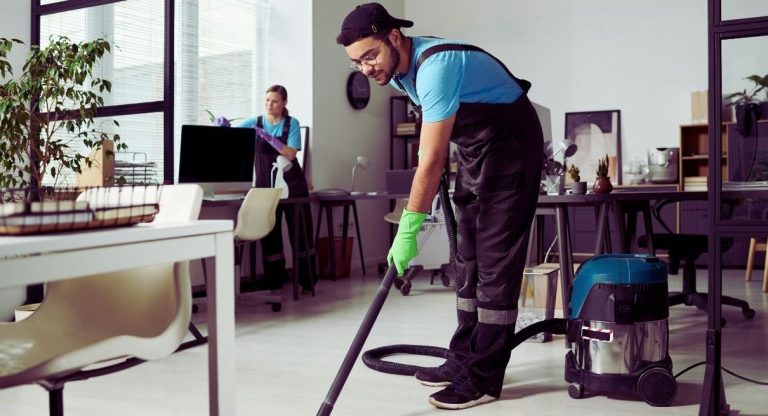 Agent d'entretien passant l'aspirateur dans un open space pendant qu'une collègue nettoie un poste de travail