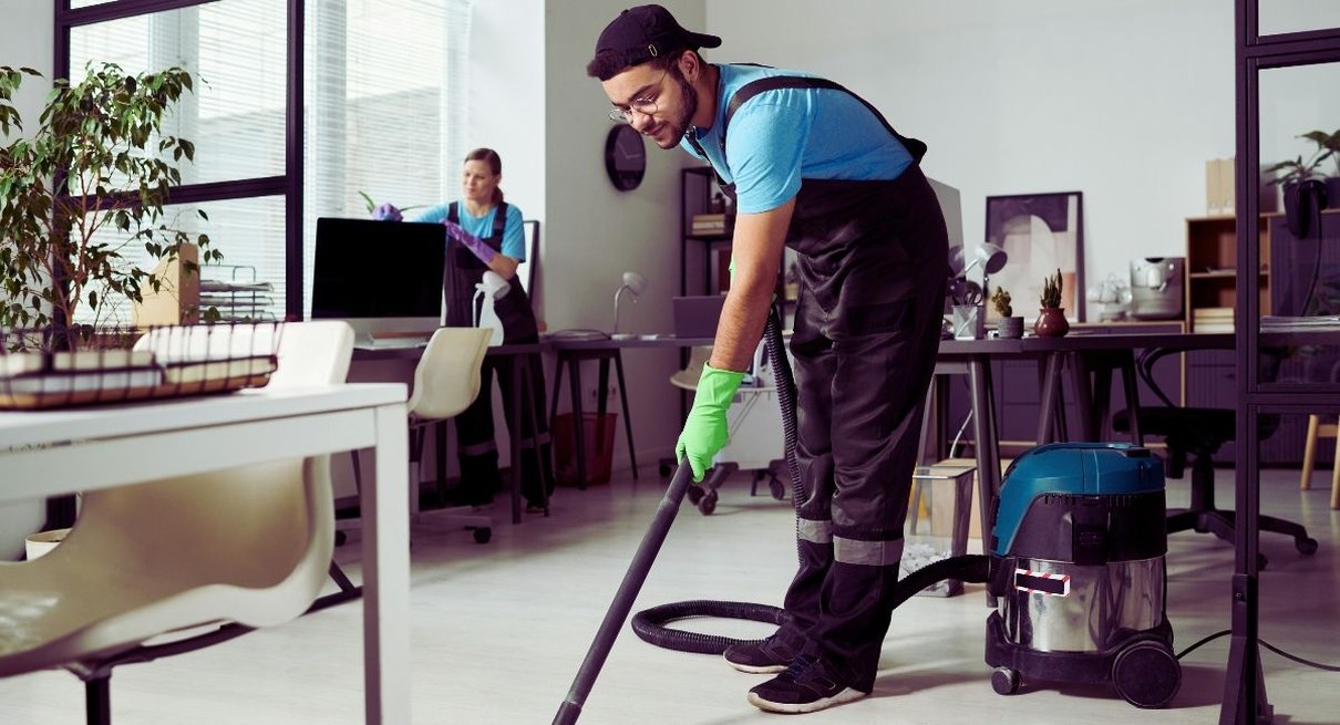 Agent d'entretien passant l'aspirateur dans un open space pendant qu'une collègue nettoie un poste de travail